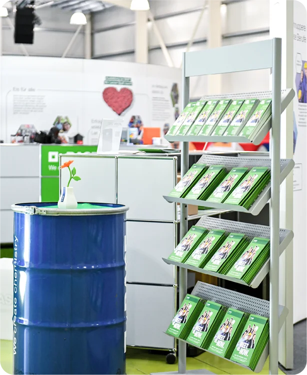 Detailansicht des Messebrandings: Graues Prospektregal mit grünen „Meet us Now“-Broschüren neben einer blauen Metalltonne mit BASF-Logo und puristischer Blumendekoration.